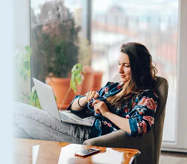 Woman sitting in an armchair with a laptop on her lap, holding a mug in a sunlit room